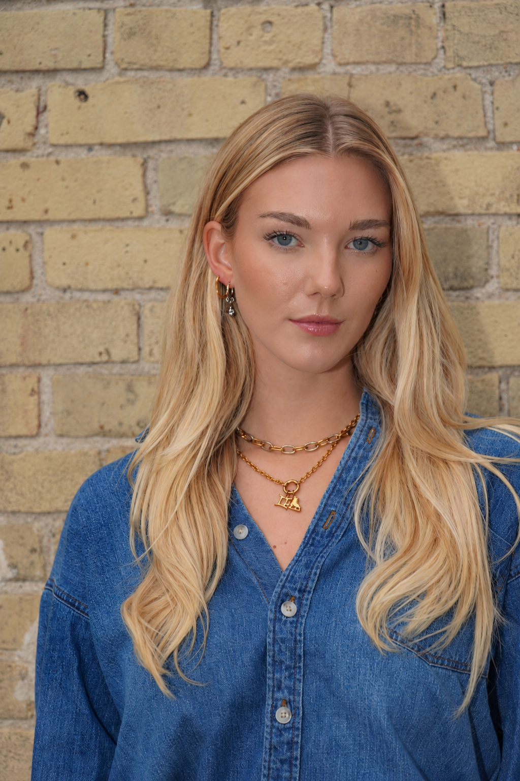 Blonde model wearing gold charm necklaces and earrings in denim against a tan brick wall